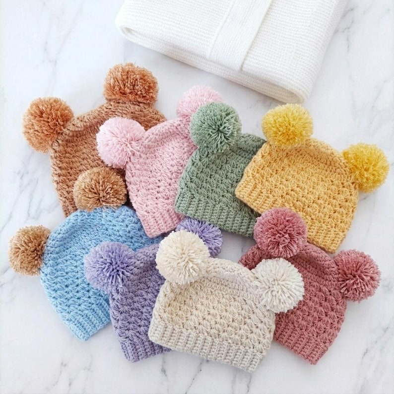 Puede incluir: Una colecci&oacute;n de ocho gorros tejidos en varios colores, incluyendo bronceado, rosa claro, verde salvia, amarillo mostaza, azul claro, lavanda, crema y rosa viejo. Cada gorro presenta dos pompones de hilo a juego en las esquinas superiores.