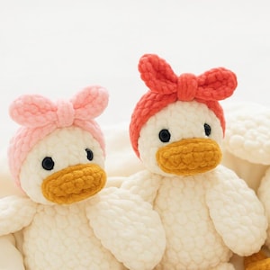 Puede incluir: Dos peluches de pato hechos a mano con ganchillo. Un pato lleva una diadema rosa con lazo, el otro un lazo rojo. Ambos tienen ojos negros y picos naranjas. Los patos son blancos con un aspecto texturizado y esponjoso.