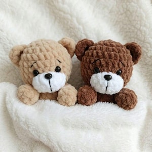 May include: Two handmade crochet teddy bears, one tan and one brown, with white muzzles and black eyes, nestled in a soft, white blanket. The bears have small, rounded ears and are made with a textured yarn.