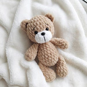 May include: A handmade, tan crochet teddy bear with a white muzzle and black eyes and nose. The bear is lying on a soft, white, fluffy blanket. The bear's arms and legs are slightly outstretched.