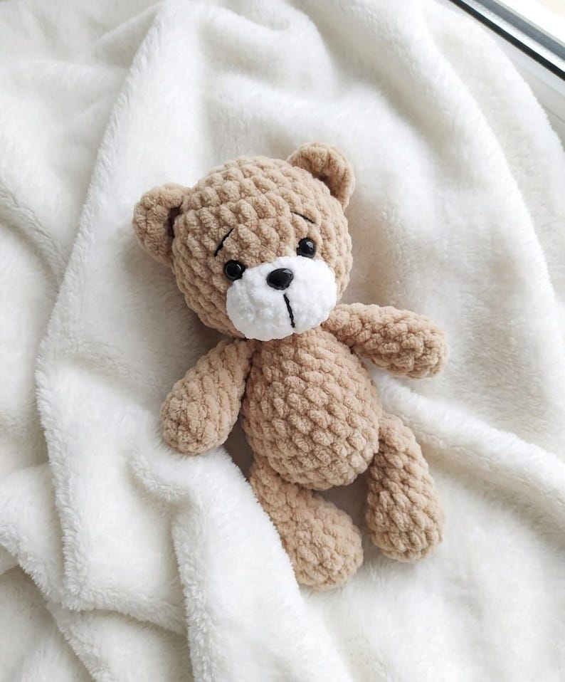 May include: A handmade, tan crochet teddy bear with a white muzzle and black eyes and nose. The bear is lying on a soft, white, fluffy blanket. The bear's arms and legs are slightly outstretched.