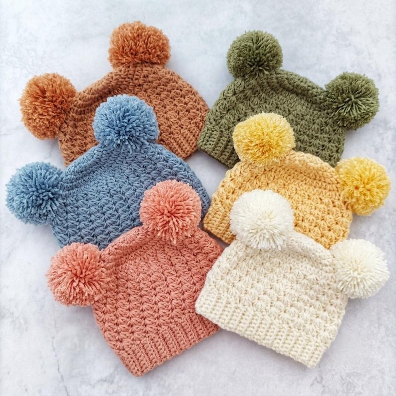 Puede incluir: Una colecci&oacute;n de cinco gorros de ganchillo en colores &oacute;xido, verde oliva, azul polvoriento, amarillo mostaza y crema. Cada gorro presenta un patr&oacute;n de punto texturizado y dos pompones de lana a juego en las esquinas superiores.
