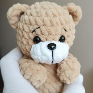 May include: A handmade, plush teddy bear in a light brown color. The bear has black eyes, a black nose, and a white muzzle. The bear is being held in a white glove. This soft toy is perfect for a baby or child.