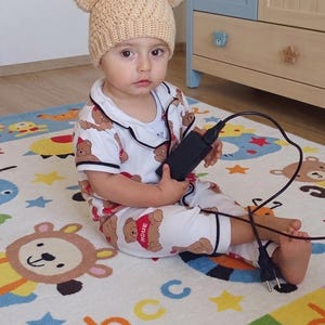 Puede incluir: Un gorro de ganchillo beige con dos pompones en la parte superior, combinado con un conjunto de pijama blanco de manga corta con estampado de osos marrones y la palabra PIQUE en un coraz&oacute;n rojo. Incluye un adaptador de corriente negro con cable.