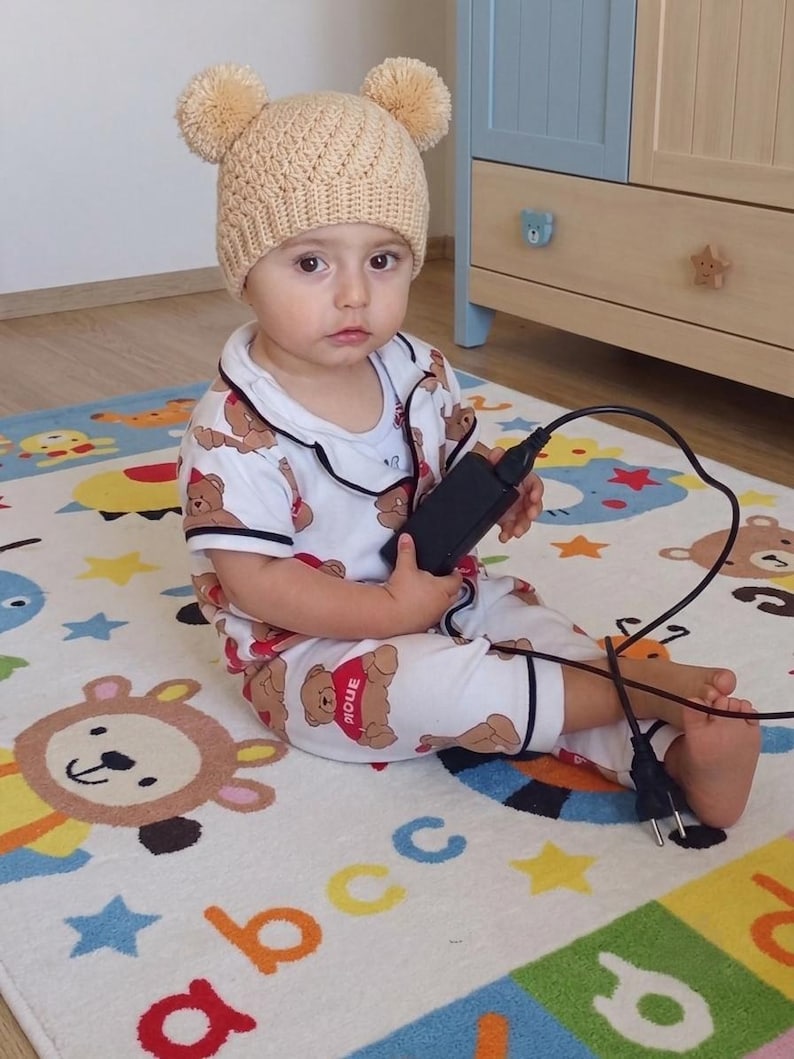 Puede incluir: Un gorro de ganchillo beige con dos pompones en la parte superior, combinado con un conjunto de pijama blanco de manga corta con estampado de osos marrones y la palabra PIQUE en un coraz&oacute;n rojo. Incluye un adaptador de corriente negro con cable.