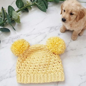 Puede incluir: Un gorro de ganchillo de color amarillo brillante con un borde acanalado y dos pompones grandes y esponjosos unidos a la parte superior. El gorro est&aacute; elaborado con un patr&oacute;n de puntos texturizado y repetitivo en todo el cuerpo principal.