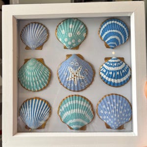 Op de afbeelding: Een vierkante witte lijst met negen individuele schelpen gerangschikt in een drie-bij-drie raster. Elke schelp is beschilderd met unieke blauwe, turquoise en witte patronen, waaronder strepen, stippen en een centraal zeesterontwerp, met goudkleurige randen.