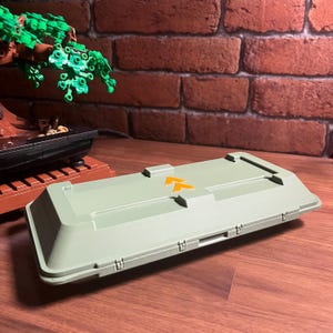 Op de afbeelding: Een lichtgroene rechthoekige opbergcontainer met een oranje pijlsymbool. De container heeft een scharnierend deksel en is gemaakt van plastic. De achtergrond omvat een bakstenen muur en een miniatuur bonsai-boom.