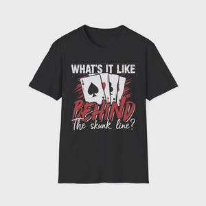 Può includere: Una t-shirt nera a maniche corte con una grafica di quattro carte da gioco. Il testo recita WHAT'S IT LIKE BEHIND The skunk line? Il design include scritte bianche e rosse con una texture invecchiata e un set di carte da gioco.