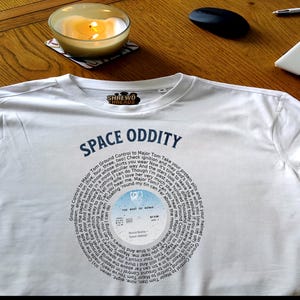 May include: White t-shirt with the text "SPACE ODDITY" and a circular graphic featuring song lyrics and a record label design. The shirt has a crew neck and the brand "SHREWD THREADS" is visible.