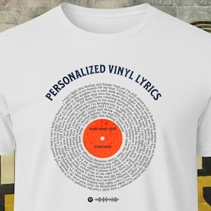 May include: White t-shirt featuring a personalized vinyl record design. The design includes the text "PERSONALIZED VINYL LYRICS" in navy blue, a circular pattern of lyrics, and a central orange circle with "BAND NAME HERE" and "SONG HERE".