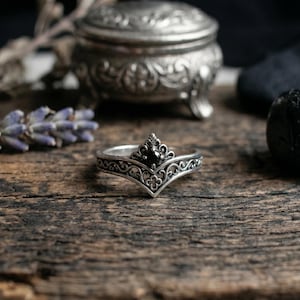 Puede incluir: Un anillo de tono plateado con una banda de filigrana intrincada y un diseño de corona en forma de V. El centro de la corona está engastado con una pequeña piedra preciosa negra redonda. El metal tiene un acabado oxidado que resalta los detalles.