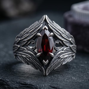 Peut inclure: Une bague en argent ornée d'une pierre précieuse rouge foncé taillée en marquise au centre. L'anneau présente des motifs de plumes texturés et détaillés qui s'étendent depuis la pierre, créant un aspect ailé avec des accents en métal oxydé.