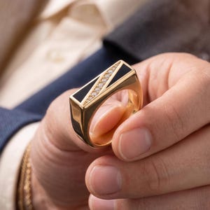 May include: A gold ring with a rectangular black onyx center and a row of small diamonds. The ring has a gold band and is held in a person's hand. The ring is a piece of jewelry.