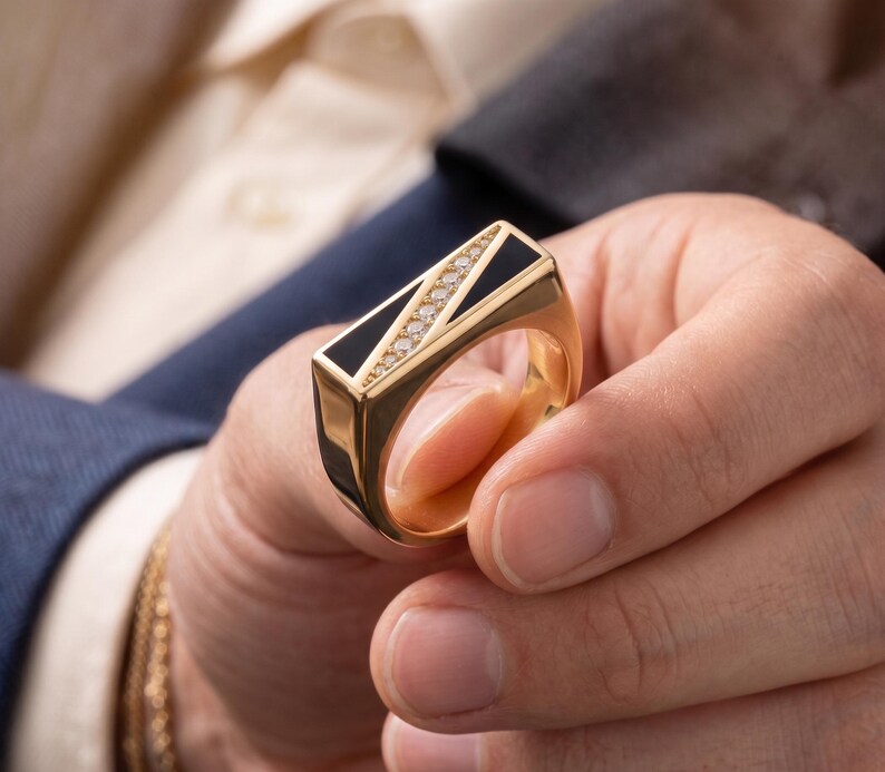 May include: A gold ring with a rectangular black onyx center and a row of small diamonds. The ring has a gold band and is held in a person's hand. The ring is a piece of jewelry.
