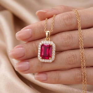 Può includere: Una collana in oro con un ciondolo rettangolare caratterizzato da una grande pietra preziosa rosso vivo al centro. La pietra rossa è circondata da un alone di piccole pietre bianche trasparenti. Il ciondolo pende da una sottile catena d'oro.