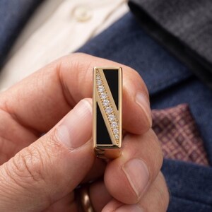 May include: A gold-toned rectangular ring with a black inlay and a diagonal row of small, clear gemstones. The ring is held in a person's hand, with a blue suit jacket and pocket square in the background.
