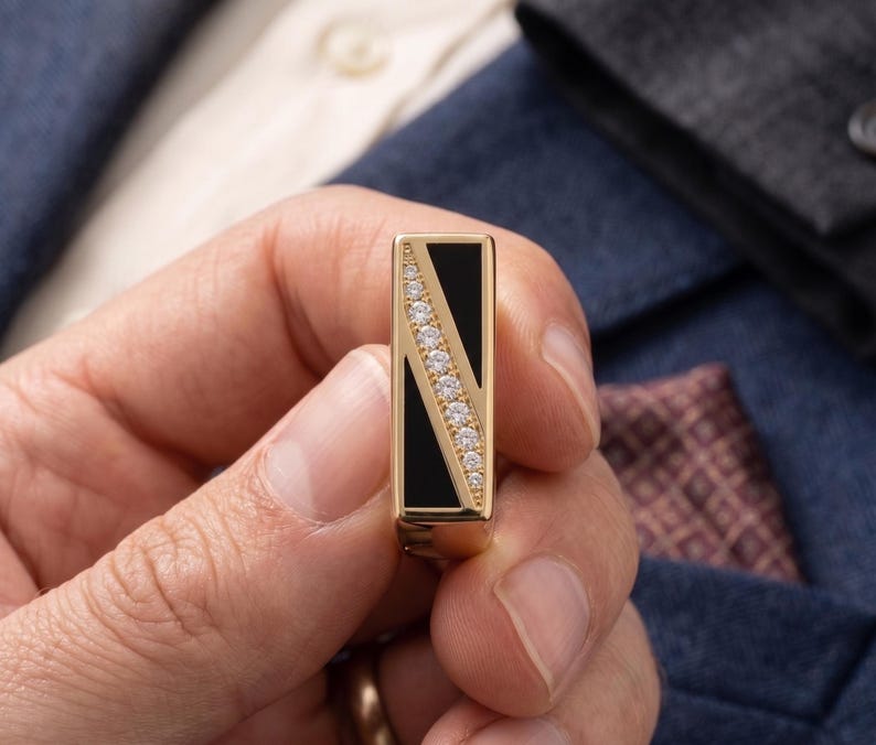 May include: A gold-toned rectangular ring with a black inlay and a diagonal row of small, clear gemstones. The ring is held in a person's hand, with a blue suit jacket and pocket square in the background.