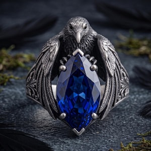 May include: A silver ring featuring a detailed raven with spread wings clutching a large, marquise-cut deep blue gemstone in its talons. The metal band displays intricate engraved feather textures and scrollwork patterns along the sides.