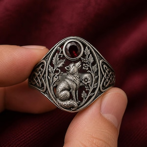May include: A silver ring featuring an oval centrepiece with a detailed relief of a wolf sitting among floral vines. A round, deep red faceted gemstone is set at the top of the oval. The sides of the ring display intricate, interlaced knotwork patterns.