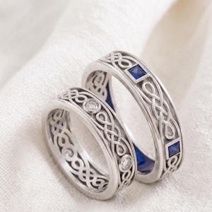 Op de afbeelding: Twee zilveren ringen met ingewikkelde Keltische knoopontwerpen. Eén ring heeft drie kleine diamanten, de andere heeft drie vierkante blauwe edelstenen. De ringen liggen op een witte stoffen achtergrond.