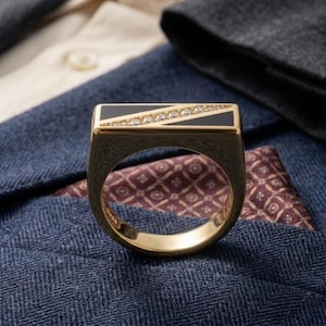 May include: A gold ring with a rectangular black onyx top, accented with a diagonal row of small diamonds. The ring is set against a backdrop of a blue blazer and a patterned pocket square.