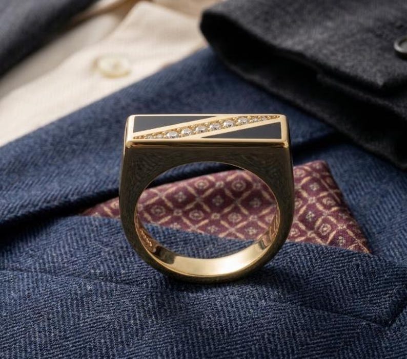 May include: A gold ring with a rectangular black onyx top, accented with a diagonal row of small diamonds. The ring is set against a backdrop of a blue blazer and a patterned pocket square.