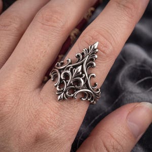 May include: A silver-toned ring featuring an elongated, pointed shield shape with intricate, symmetrical scrollwork and filigree detailing. The metal surface has darkened recessed areas to highlight the raised, polished decorative patterns.