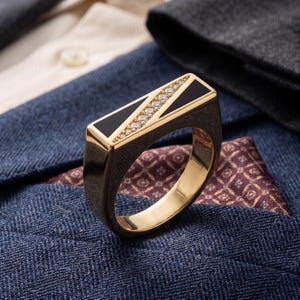 May include: A gold ring with a rectangular black onyx inlay and a row of small diamonds. The ring is set against a backdrop of a blue blazer, a patterned pocket square, and a white shirt.