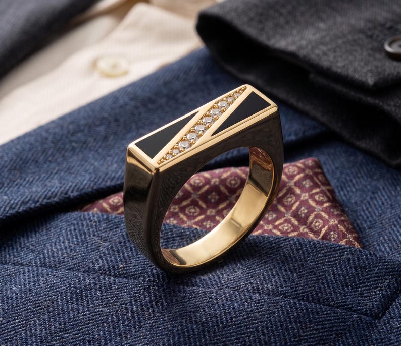 May include: A gold ring with a rectangular black onyx inlay and a row of small diamonds. The ring is set against a backdrop of a blue blazer, a patterned pocket square, and a white shirt.