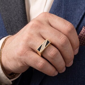 May include: A gold ring with a rectangular black onyx inlay and a row of diamonds. The ring is on a hand, with a gold bracelet visible on the wrist. The person is wearing a blue blazer with a patterned pocket square.