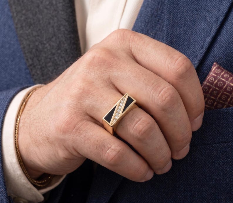May include: A gold ring with a rectangular black onyx inlay and a row of diamonds. The ring is on a hand, with a gold bracelet visible on the wrist. The person is wearing a blue blazer with a patterned pocket square.