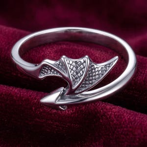 May include: A silver-toned open ring featuring a detailed dragon wing design on one end and a pointed arrow-like tail on the other. The metal surface has a polished finish with textured scale patterns on the wing, creating a wrap-around effect.