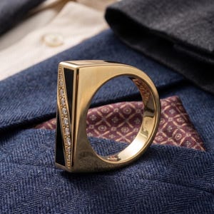 May include: A gold ring with a black rectangular inlay and a row of small diamonds. The ring is set against a backdrop of blue and cream-colored fabric.