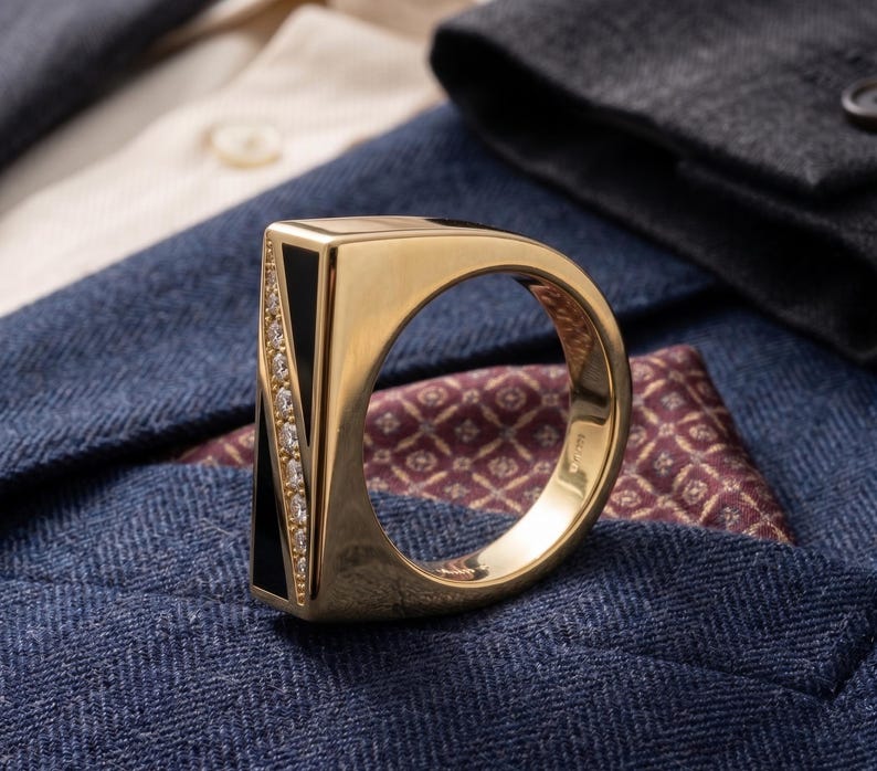 May include: A gold ring with a black rectangular inlay and a row of small diamonds. The ring is set against a backdrop of blue and cream-colored fabric.