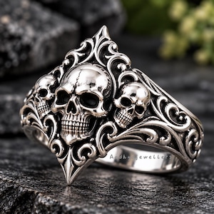May include: A silver-toned ring featuring a large central skull flanked by two smaller skulls, all integrated into an ornate, raised scrollwork design. The metal surface is polished with dark recessed areas. The text Aadhyajewellers is visible.