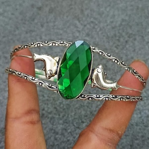 May include: A silver-toned cuff bracelet featuring a large, oval-shaped, faceted green gemstone at the center. The band is decorated with a textured, wavy pattern and includes two silver-toned dolphin figures positioned on either side of the stone.