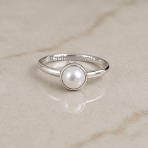 May include: A silver-toned ring featuring a single, round, white pearl set in a smooth bezel setting. The band is polished and metallic. The text Aadhyajewelrs is visible in a light grey font across the centre of the ring band.