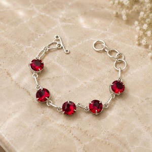 May include: A silver-toned bracelet featuring five round, faceted deep red stones set in prong settings. The chain consists of delicate silver links, ending with a toggle clasp on one side and a series of circular adjustment rings on the other.