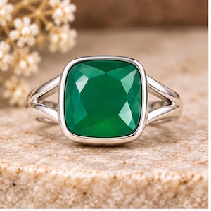 May include: A silver ring featuring a large, square-cut green gemstone with rounded corners. The stone is set in a smooth metal bezel, and the band consists of two split, polished silver shanks that meet at the sides of the central setting.