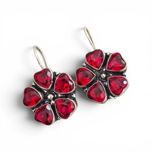 Puede incluir: Un par de pendientes colgantes de plata con un diseño en forma de flor. Cada pendiente consta de cinco piedras rojas facetadas en forma de corazón, dispuestas en un patrón circular alrededor de un punto central de plata y un gancho.