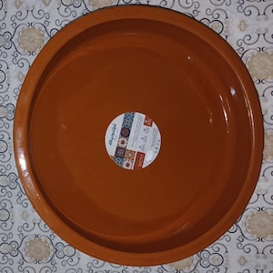 Può includere: Un grande piatto da portata rotondo color terracotta. Il piatto ha un design ampio e poco profondo con un bordo leggermente rialzato. Un adesivo circolare con testo arabo e motivi decorativi è al centro. Il piatto è probabilmente in ceramica o terracotta.
