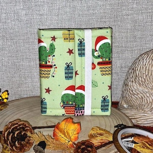 May include: A green fabric wallet with a Christmas cactus pattern, including Santa hats, fairy lights, and gift boxes. The wallet is held closed with a white elastic band. The background features autumn leaves and a decorative rabbit statue.