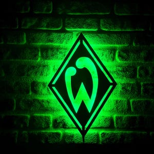 Puede incluir: Logotipo verde brillante del Werder Bremen, un diamante con una 'W' en su interior, sobre una pared de ladrillos. El logotipo brilla, contrastando con el fondo oscuro. Se aprecia el trabajo de los ladrillos.