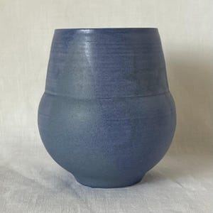 May include: A blue ceramic vase with a rounded base and a flared top. The vase has a textured surface and a matte finish. The color is a muted, dusty blue, adding a touch of rustic charm. The vase is suitable for displaying flowers or as a decorative item.
