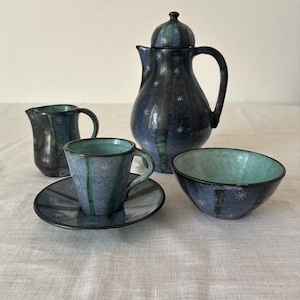 May include: A ceramic tea set featuring a tall lidded teapot, a small creamer, a cup on a saucer, and a bowl. Each piece has a dark blue and teal glaze with a speckled finish and small white star-like patterns scattered across the surfaces.