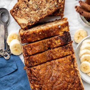 Puede incluir: Vista aérea de pan de plátano en rodajas con nueces. También hay plátanos en rodajas, cucharas medidoras y palitos de canela. El pan tiene una corteza dorada y un interior húmedo, perfecto para un refrigerio o desayuno.
