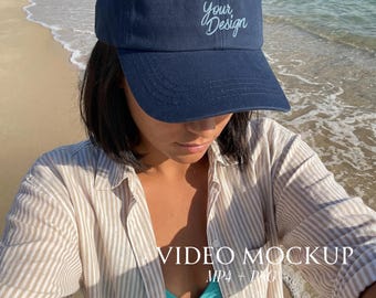 May include: A navy blue baseball cap with the text "Your Design" in a script font. The image also includes the text "VIDEO MOCKUP MP4 + PNG". The cap is worn near the ocean.