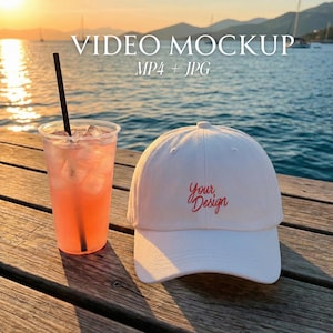 May include: A white baseball cap with the text "Your Design" in red script sits next to a clear plastic cup filled with a pink beverage and a black straw. The background shows a body of water and the text "VIDEO MOCKUP MP4 + JPG".