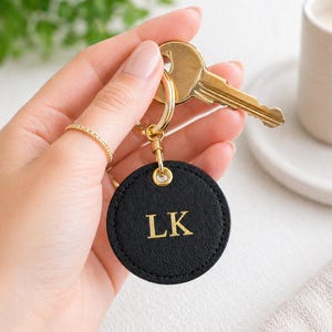 May include: A round, black leather keyring with gold-coloured hardware and the gold initials "LK". The keyring is attached to a set of keys. A candle and greenery are visible in the background.